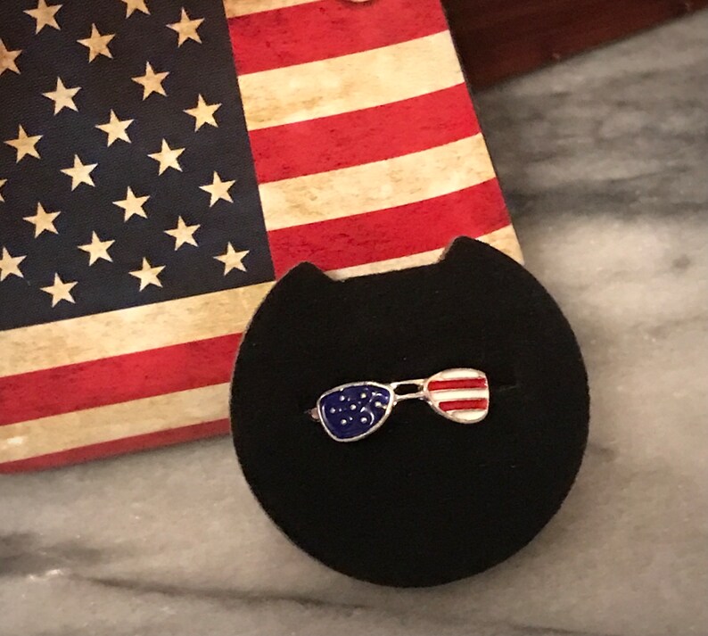 May include: A silver ring featuring an enamel design of sunglasses in blue and red. The sunglasses are decorated with stars and stripes, reminiscent of the Union Jack.