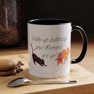 Peut inclure: Un mug en céramique blanc avec un intérieur et une anse noirs, portant le texte noir 'Wake up buttercup your Demons are up!' accompagné d'une illustration de chat noir et de deux petits monstres colorés avec des motifs de cœur.
