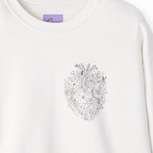 Peut inclure: Un sweat-shirt à col rond blanc cassé avec une illustration au trait noir sur la poitrine. Le motif représente une forme de cœur anatomique composée de diverses fleurs, feuilles et petites étoiles, dans un style détaillé et délicat.