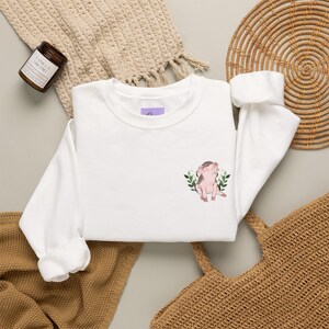 Peut inclure: Un sweat-shirt blanc à manches longues avec un petit motif brodé sur la poitrine gauche. Le dessin représente un porcelet rose clair assis, entouré d'un groupe de feuilles vertes et de petites fleurs roses, dans un style doux et délicat.