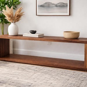 May include: A dark brown wooden media console table with an open shelf design. The table is against a white wall. A white vase with dried flowers, a black bowl, books, and a woven basket are on top. The table is approximately 150 cm long.