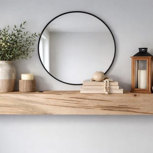 May include: A contemporary interior design display featuring a wooden floating shelf with a black-framed circular mirror. The shelf is adorned with a vase of greenery, a candle, books, and decorative objects. A wooden lantern and a vase with dried flowers are also present.