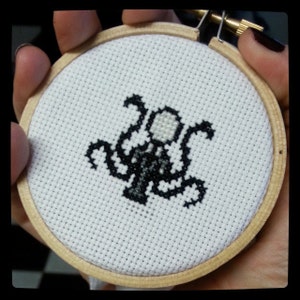 May include: A black and white cross-stitch embroidery of a creature with multiple arms and a round head. The embroidery is framed in a wooden hoop.