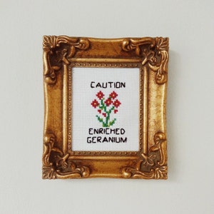 May include: A framed cross-stitch with a gold frame. The cross-stitch reads "CAUTION ENRICHED GERANIUM" and depicts a red flower with green leaves.