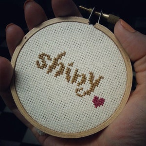 May include: A close-up of a hand holding a small, round embroidery hoop with the word "Shiny" stitched in gold thread and a small red heart stitched below it.