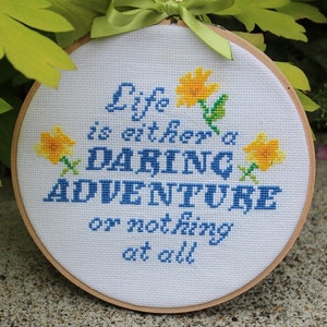 May include: A white embroidery hoop with a blue and yellow cross-stitch design. The design reads "Life is either a daring adventure or nothing at all".