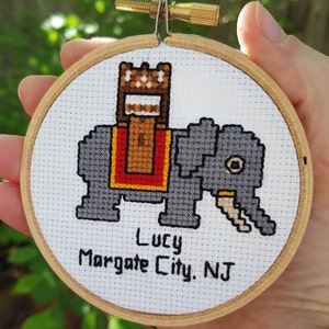 May include: A white embroidery hoop with a gray elephant carrying a brown castle-shaped object. The embroidery reads "Lucy Margate City, NJ".