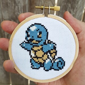 May include: A blue and brown cross-stitch embroidery of a Squirtle Pokemon character in a wooden hoop.
