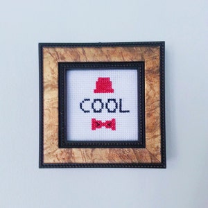 May include: A framed cross-stitch with a red hat, a bow tie, and the word "COOL" in black letters.