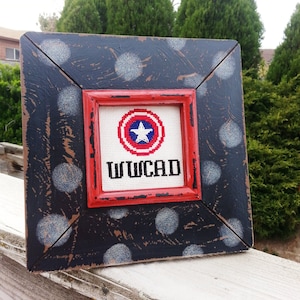 May include: A framed cross-stitch featuring a red, white and blue American flag design and the text "WWCAD" in white. The frame is black with white polka dots.