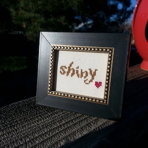 May include: A framed cross-stitch with the word "shiny" in brown thread and a small red heart in the corner.