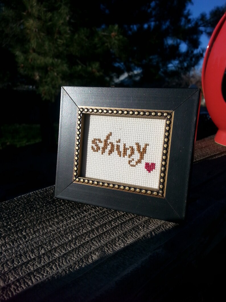 May include: A framed cross-stitch with the word "shiny" in brown thread and a small red heart in the corner.