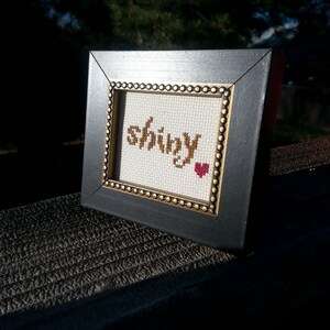 May include: A small black frame with a gold trim surrounds a white cross-stitch piece that reads "shiny" with a red heart.