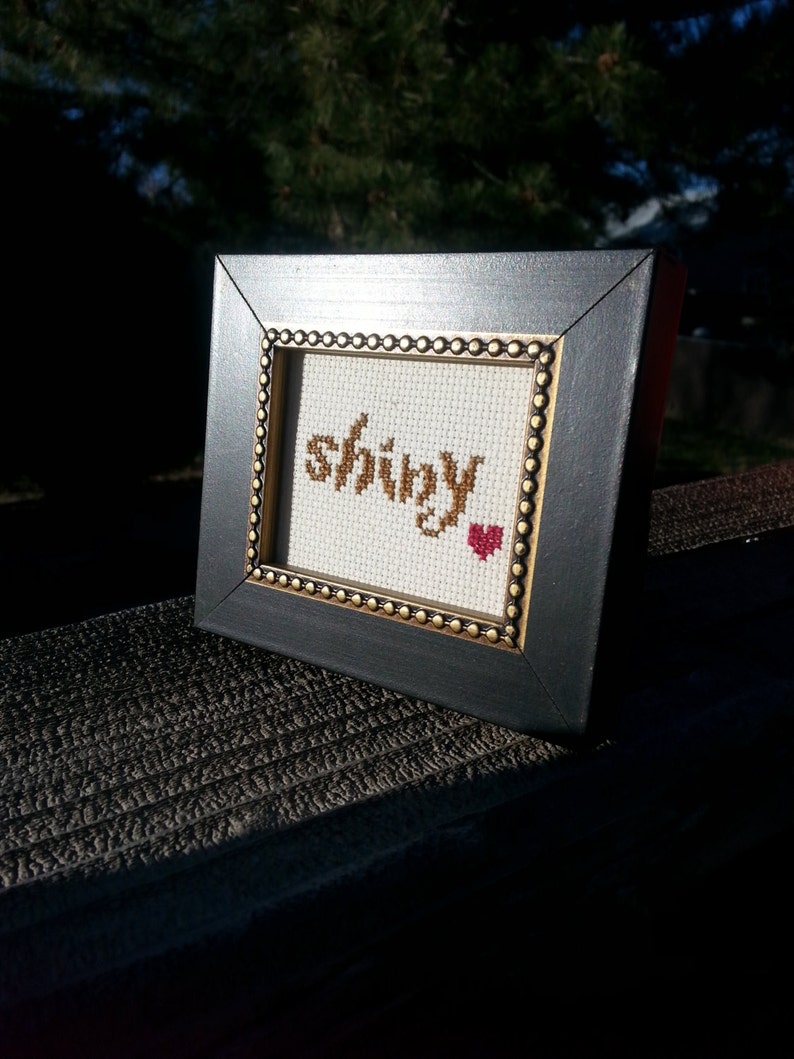 May include: A small black frame with a gold trim surrounds a white cross-stitch piece that reads "shiny" with a red heart.