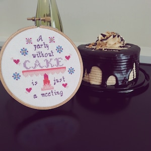 May include: A cross-stitch embroidery hoop with the saying "A party without cake is just a meeting". The hoop is on a black surface next to a chocolate cake with peanut butter frosting.