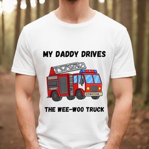 Peut inclure: T-shirt blanc avec le texte "MY DADDY DRIVES THE WEE-WOO TRUCK" au-dessus d'une illustration de camion de pompiers rouge avec une échelle, des feux bleus et des pneus noirs. Le t-shirt est à manches courtes.