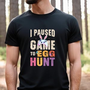 Könnte beinhalten: Schwarzes T-Shirt mit dem Text "I PAUSED GAME TO EGG HUNT". Das Design zeigt einen Gamecontroller mit Hasenohren und farbigem Text. Das T-Shirt hat einen Rundhalsausschnitt.
