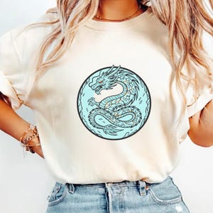 Puede incluir: Camiseta color crema con un diseño circular que presenta una ilustración de un dragón azul. El dragón tiene detalles en naranja y está sobre un fondo azul claro dentro de un círculo negro. La camiseta tiene mangas cortas.