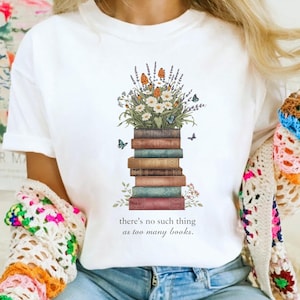 May include: White t-shirt with a design of a stack of books topped with flowers and butterflies. The text reads "there's no such thing as too many books". The shirt is made of a soft material.