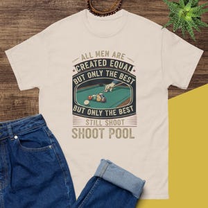 May include: Cream-colored t-shirt with a graphic featuring a pool table and the text "ALL MEN ARE CREATED EQUAL BUT ONLY THE BEST STILL SHOOT POOL." The design includes a hand holding a pool cue.
