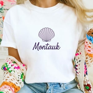 May include: White t-shirt featuring a purple seashell graphic and the word "Montauk" in a matching purple script. The shirt is made of a soft material and is suitable for casual wear. The overall theme is coastal and beach-inspired.