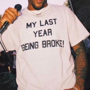 May include: A white t-shirt with black text that reads "My Last Year Being Broke!"