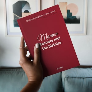 Peut inclure: Un livre bordeaux intitulé "Maman Raconte moi ton histoire" en écriture blanche. Le livre est tenu dans une main avec une bague argentée et du vernis à ongles blanc. La couverture du livre comprend également le texte "Un livre à compléter à coeur ouvert".