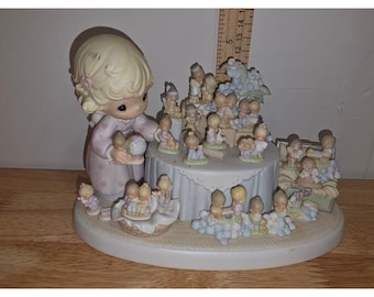 Może przedstawiać: Zestaw figurek porcelanowych przedstawiający dużą postać dziecka w różowym stroju w kropki, trzymającą mały globus, otoczoną licznymi mniejszymi figurkami dzieci na okrągłej podstawie. Drewniana linijka wskazuje wysokość 12,7 cm.