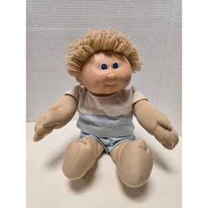 May include: A soft-bodied doll with a round vinyl face, dimpled cheeks, and bright blue eyes. The doll has short, textured blonde yarn hair and is wearing a two-toned outfit consisting of a white short-sleeved top and light blue shorts.
