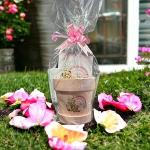 May include: A pink flower pot with a teddy bear design, wrapped in clear cellophane and tied with a pink ribbon. The pot has the text "Mrs. Smith" and "Thank you" on it. The pot is filled with small items.