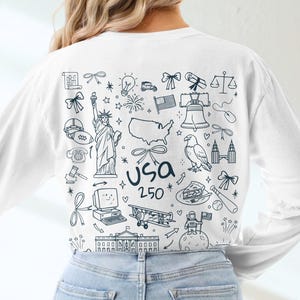 May include: A white long-sleeved top with a USA 250 graphic. The design includes American icons such as the Statue of Liberty, a map of the USA, the Liberty Bell, and an eagle, all in a doodle style.