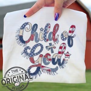 Może przedstawiać: Biała koszulka z napisem "Child of God" w kolorach czerwonym, białym i niebieskim, ze wzorem amerykańskiej flagi w stylu vintage. Koszulka ma również czarną pieczęć z napisem "The Original Designer".