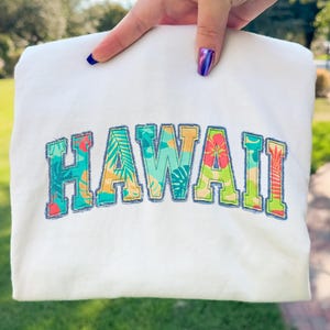 以下が含まれることがあります： 前面に「HAWAII」の文字が刺繍された白いスウェットシャツ。文字は、緑、青、オレンジ、黄色のトロピカルな花と葉の模様で埋められています。スウェットシャツは人が持っています。
