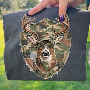 Könnte beinhalten: Dunkelgraues Sweatshirt mit einem aufgestickten Aufnäher eines Hirsches mit Sonnenbrille und Camouflage-Hut. Das Reh ist vor einem Camouflage-Schild platziert.