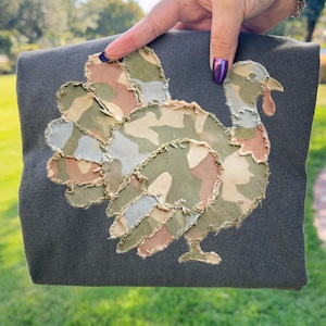 Könnte beinhalten: Dunkelgraues Stoffstück mit einem Truthahn-Applikation. Der Truthahn besteht aus Camouflage-Stoff in Grün-, Braun- und Beigetönen, mit rotem Wamm und Schnabel. Die Applikation hat einen ausgefransten Rand. Der Artikel wird vor einem grünen Hintergrund gehalten.