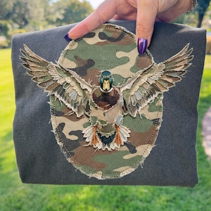 Può includere: Una maglietta grigio scuro con un'applicazione dettagliata di un'anatra con le ali spiegate. L'anatra è posta su un ovale mimetico, con le ali e il corpo dell'anatra in tonalità marroni, verdi e beige.