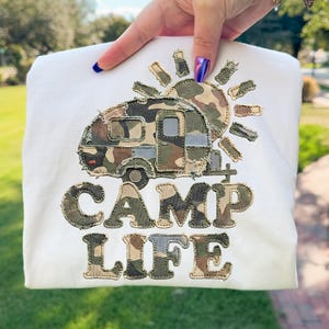 May include: A white sweatshirt featuring a camper design with a camouflage pattern and the words "CAMP LIFE" in a matching camouflage style. The sun is also designed with a camouflage pattern.