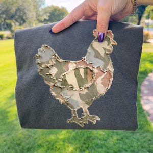 May include: A dark gray tote bag featuring a camouflage chicken applique. The chicken design incorporates shades of green, brown, and beige. The bag is held up against a backdrop of green grass and a blue sky.