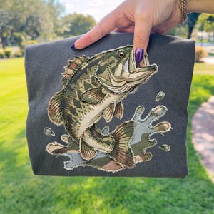 Może przedstawiać: Ciemnoszara bluza z haftem przedstawiającym bass wielkogębowy wyskakujący z wody. Ryba ma wzór kamuflażu w odcieniach zieleni, brązu i beżu. Rozbryzg wody jest również w kamuflażu. Bluza jest trzymana przez osobę.