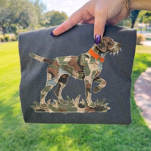 Puede incluir: Artículo de tela gris oscuro con un diseño de perro bordado. El perro está representado en un patrón de camuflaje con un collar naranja, de pie sobre hierba verde. El fondo es una escena borrosa al aire libre con hierba verde y árboles.