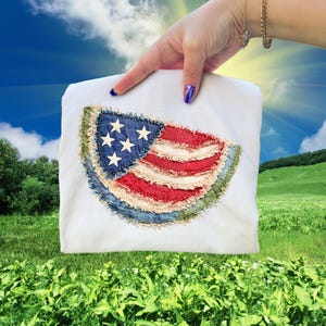 May include: White t-shirt featuring a distressed American flag design. The flag is made of blue denim with white stars, and red and white striped fabric. The shirt is held against a backdrop of a sunny sky and green field.