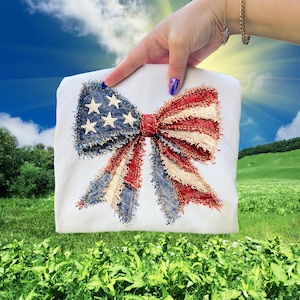 May include: White t-shirt featuring a large bow design in red, white, and blue, reminiscent of the American flag. The bow has a denim-like texture with white stars on the blue section. The design is centered on the shirt.
