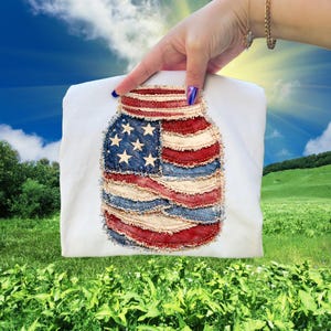 May include: A white t-shirt with a patriotic design of a mason jar with the American flag. The jar design is made of red, white, and blue fabric with a distressed, frayed edge. The shirt is held up against a backdrop of a sunny sky and green field.