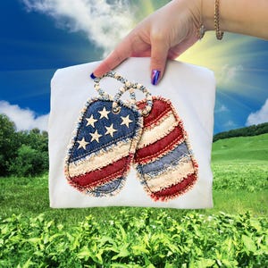 May include: A white t-shirt featuring two decorative dog tags. One tag has a blue denim background with white stars and red and white stripes. The other tag has a red and white striped design. Both tags have frayed edges.