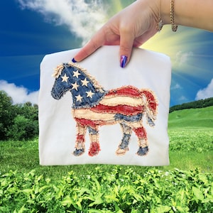 Può includere: Una maglietta bianca con un motivo a cavallo applicato. Il cavallo è decorato con la bandiera americana, con strisce rosse e bianche per il corpo e una testa blu con stelle bianche. La maglietta è mostrata su uno sfondo di campo verde e cielo blu.
