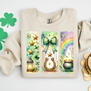 Puede incluir: Sudadera beige con un diseño del Día de San Patricio. El diseño incluye una linterna, estrellas, tréboles, una herradura, un lazo verde, un arcoíris y una olla de oro. La obra de arte es de estilo acuarela.