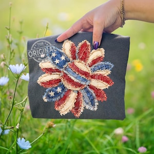 May include: A dark gray t-shirt featuring a patriotic sunflower design. The flower petals are made of red, white, and blue fabric, with stars on the blue petals. The text "THE ORIGINAL DESIGN" is printed on the shirt.