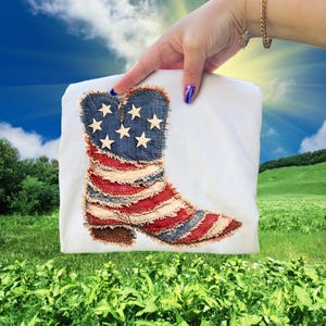 May include: White t-shirt featuring a patriotic design of a cowboy boot. The boot is made of denim and red and white fabric, with a blue denim top adorned with white stars. The design evokes the American flag.
