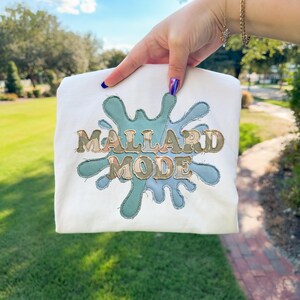 May include: White sweatshirt with a teal and green splash design. The words "MALLARD MODE" are embroidered in a vintage style. The sweatshirt is held up against a background of green grass and trees.