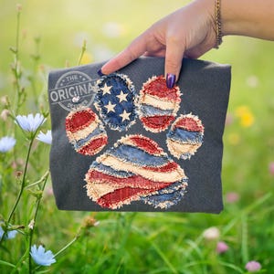 May include: Dark grey t-shirt with a paw print design made from red, white, and blue fabric patches, mimicking the American flag. The design incorporates stars and stripes. The text "THE ORIGINAL DESIGNER" is printed on the upper left corner.